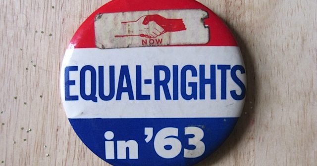 Red, white, and blue stripped button laying on a wodden surface. at the top of the button, there is a sticker with two hands that says