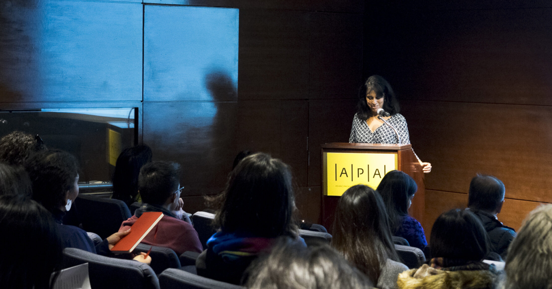 A photograph of Crystal Parikh standing at a podium that reads A/P/A Institute. An audience sits facing her, with their backs towards the camera.