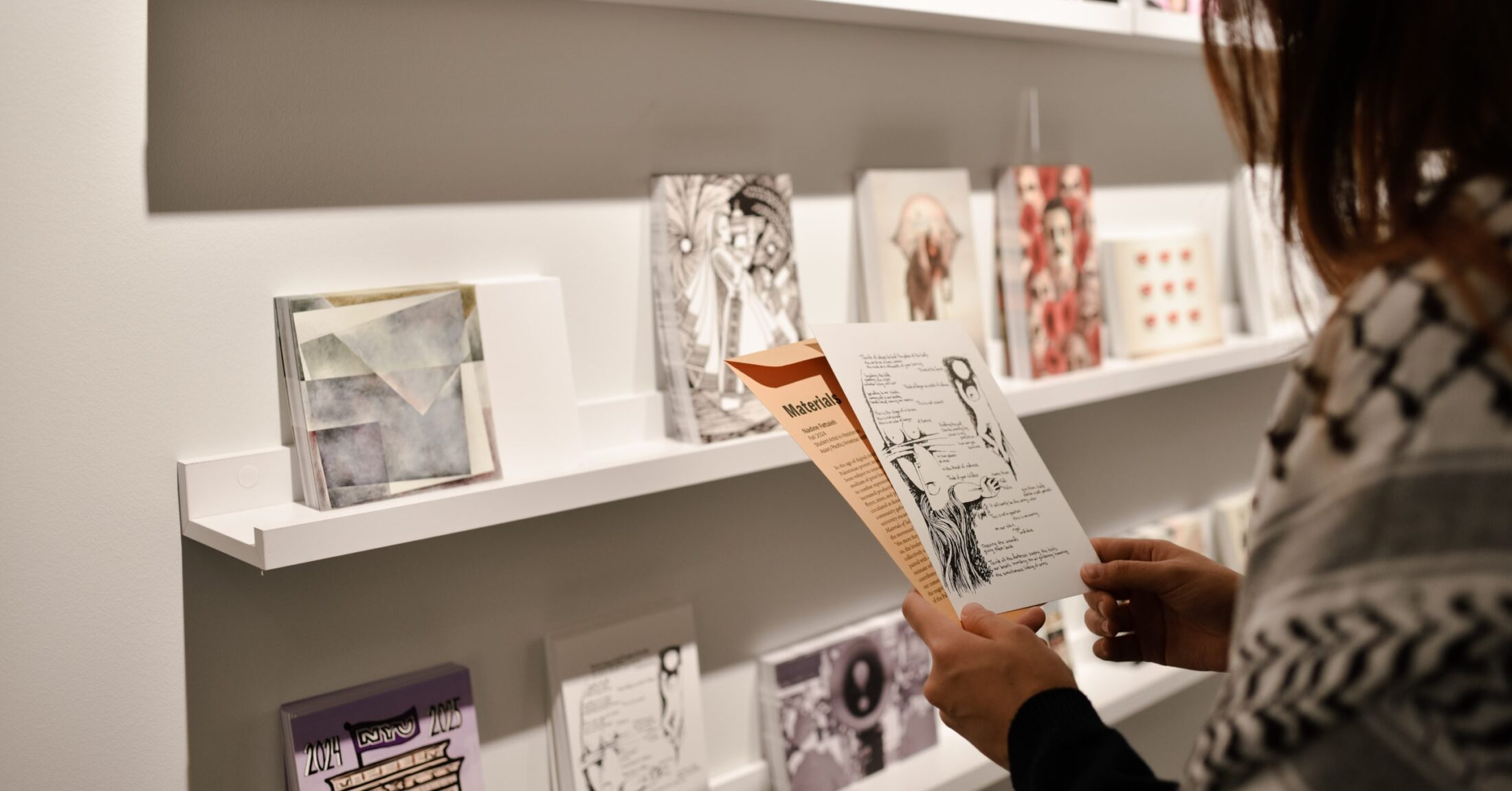 A photograph of a visitor holding a postcard in the exhibition Materials of Solidarity. Postcards placed on white shelves are visible in front of them.