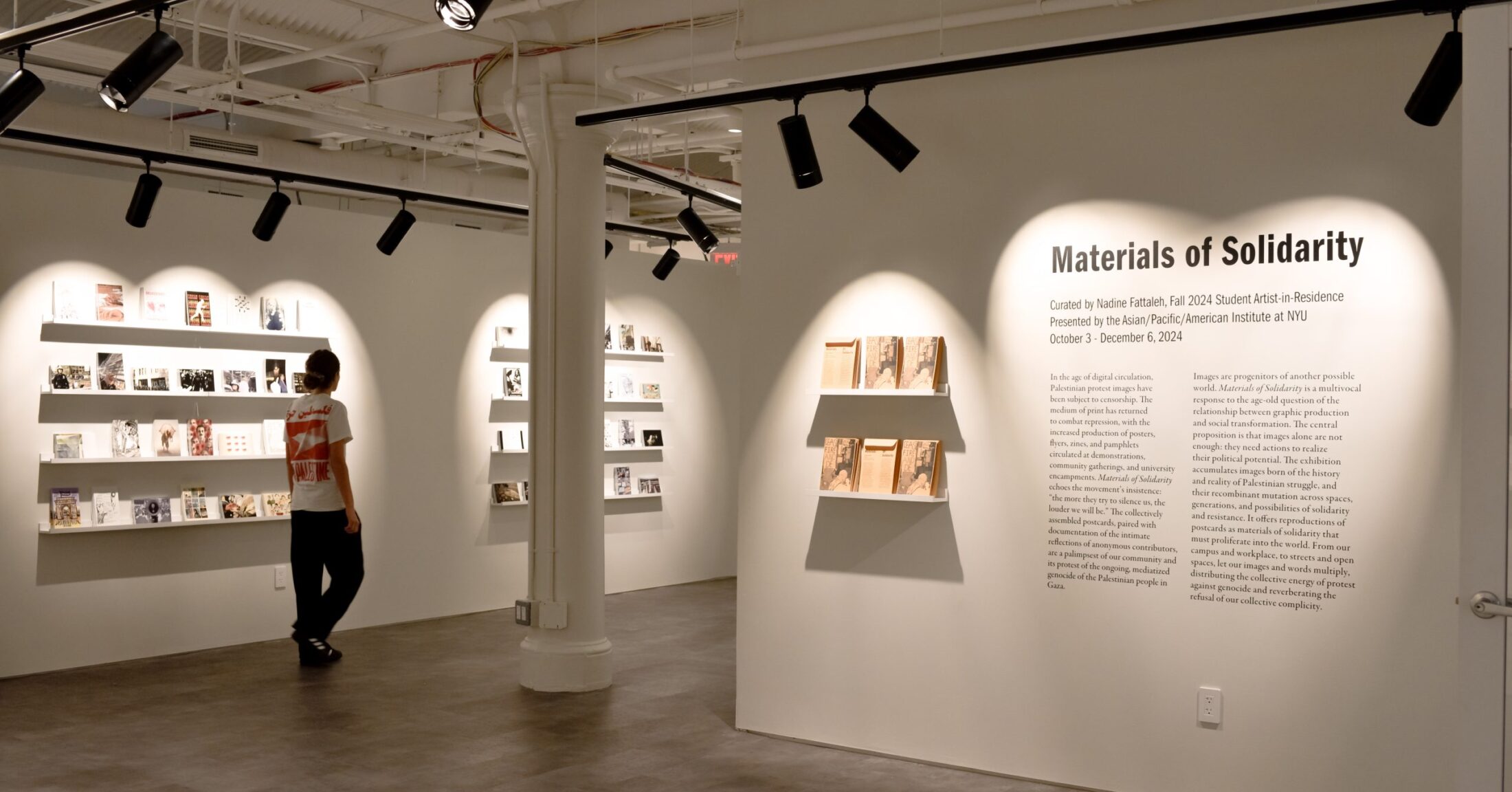 A photograph of the exhibition Materials of Solidarity. It features vinyl text describing the exhibition, and white shelves on which postcards and exhibition materials are placed.