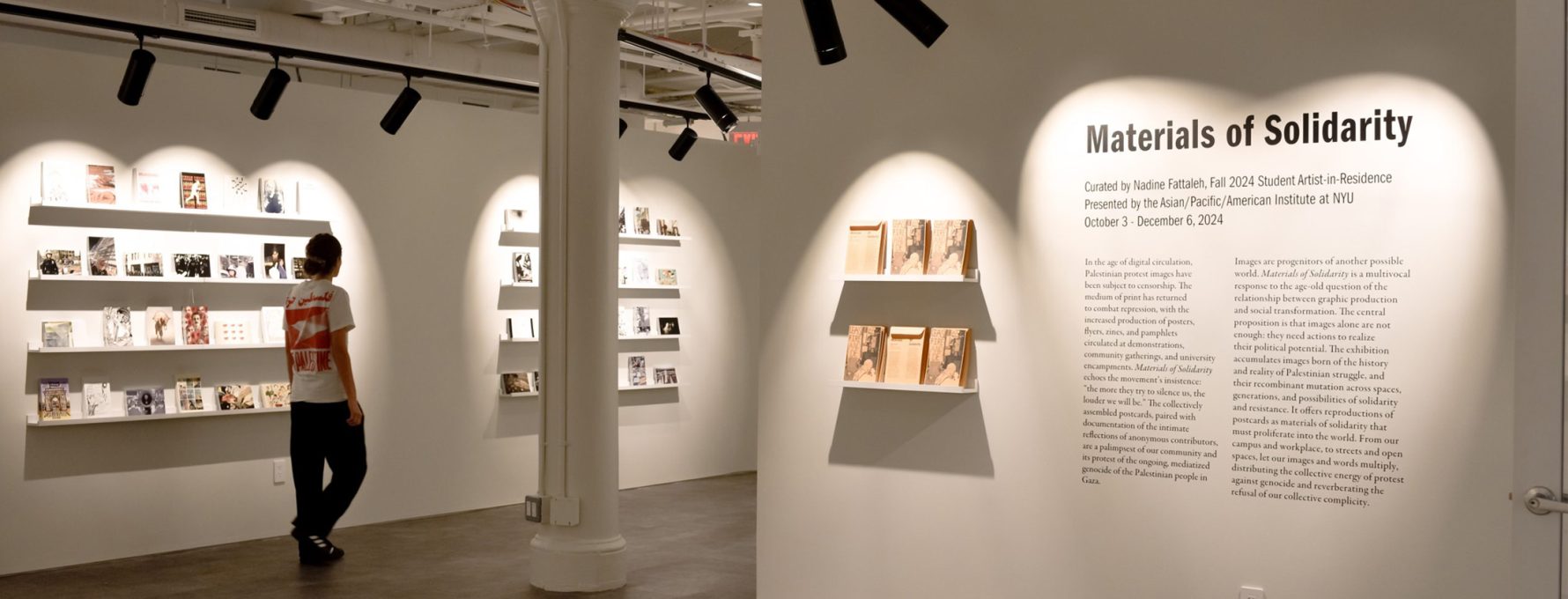 A photograph of the exhibition Materials of Solidarity. It features vinyl text describing the exhibition, and white shelves on which postcards and exhibition materials are placed.
