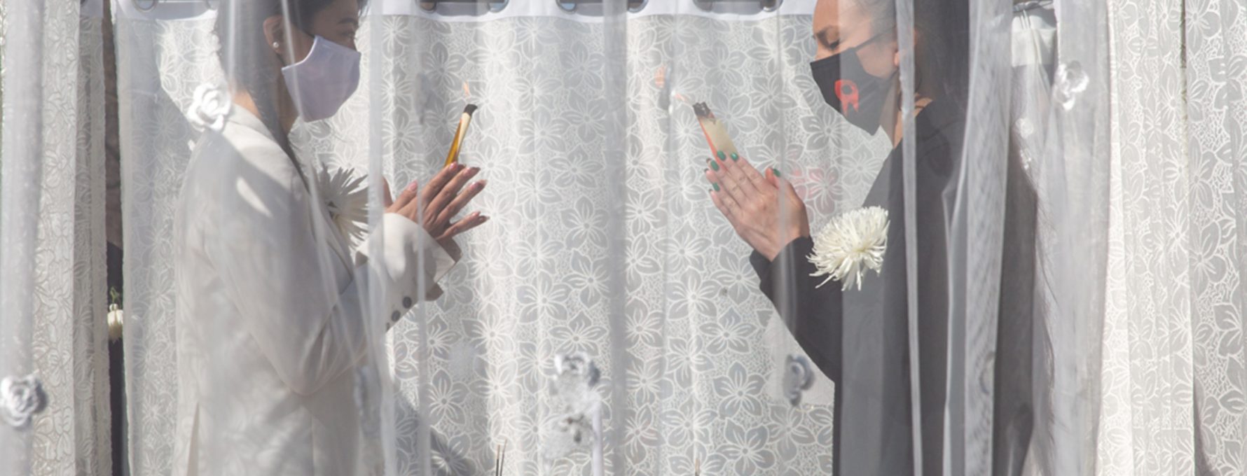 A photograph of a Red Canary Song altar performance, which took place in Washington Square Park in 2022. RCS members stand in front of an altar, behind a lace curtain, and facing each other. Their hands are in pray position and they hold incense.