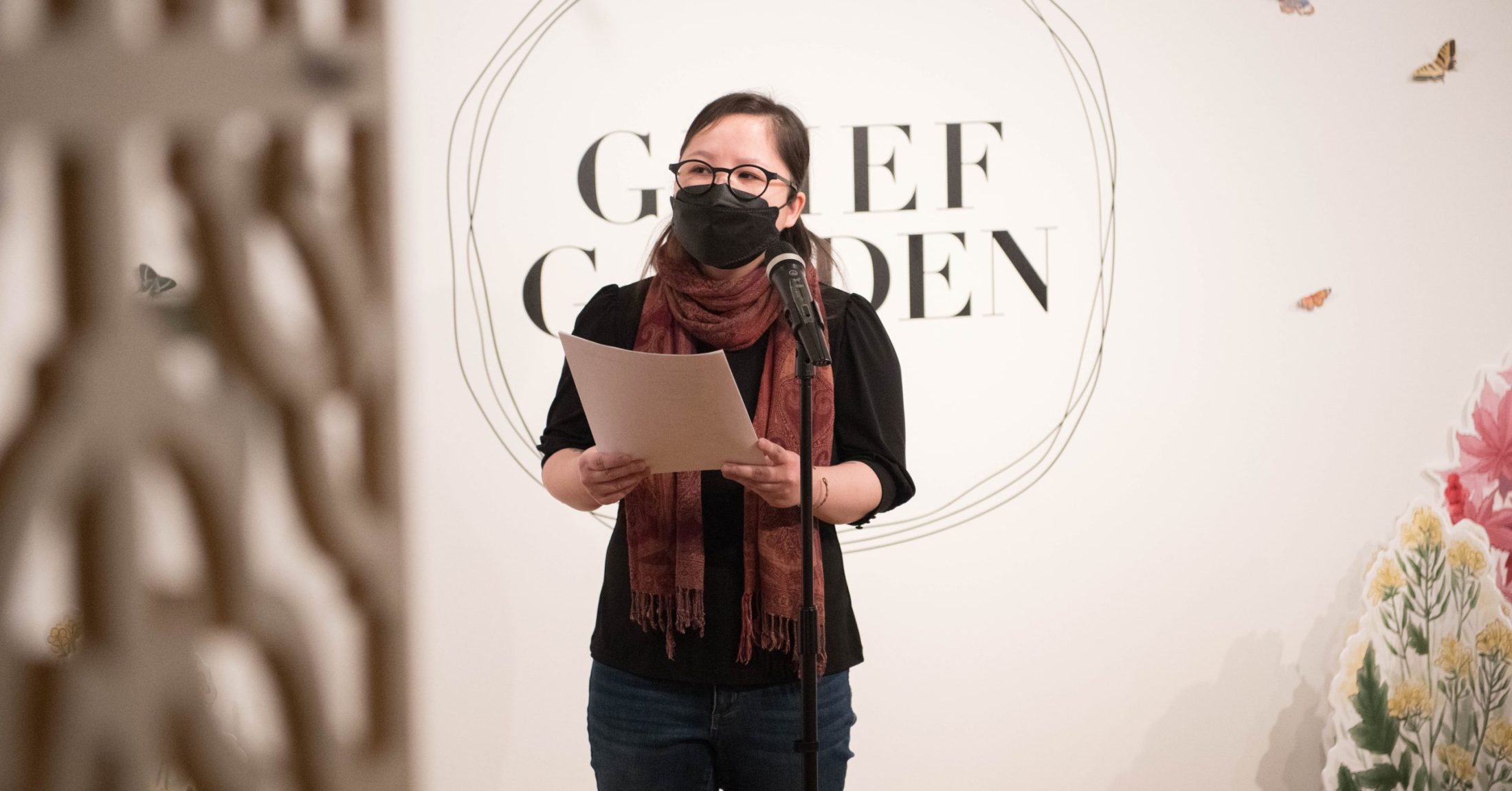 Photograph of Khaty Xiong. She stands in front of a mic reading, and holds paper in her hand.