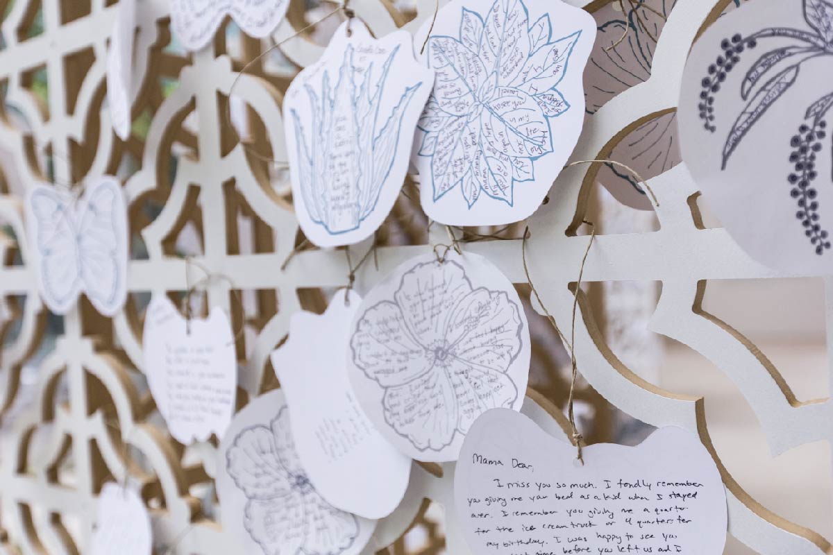 Photo of piece in art installation. Small paper cut-outs with written words, as well as drawings of flora and fauna, hang from an ornate wooden screen.