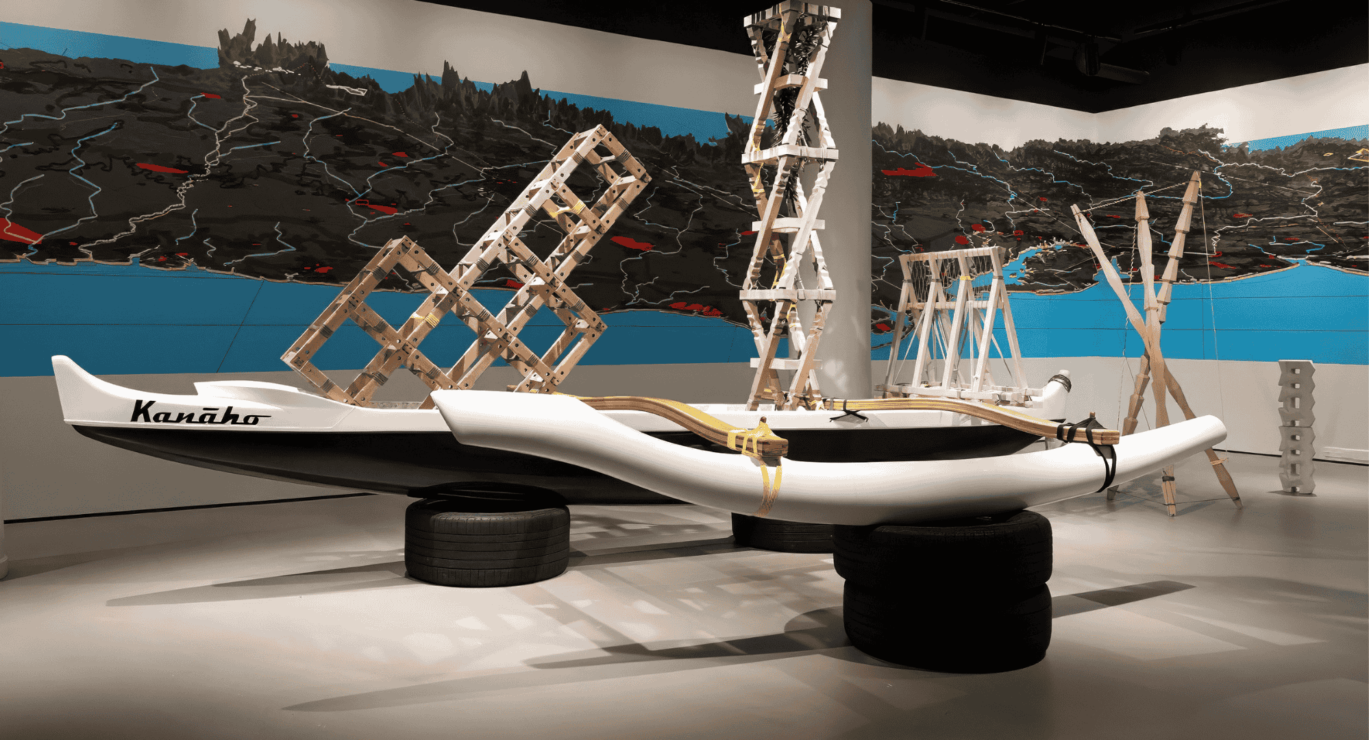 A photograph of the installation, Hawai'i is not the US but it is your Future, by Sean Connelly. The photograph features sculpture and mapping work by Connelly, in addition to a Hawaiian outrigger canoe.