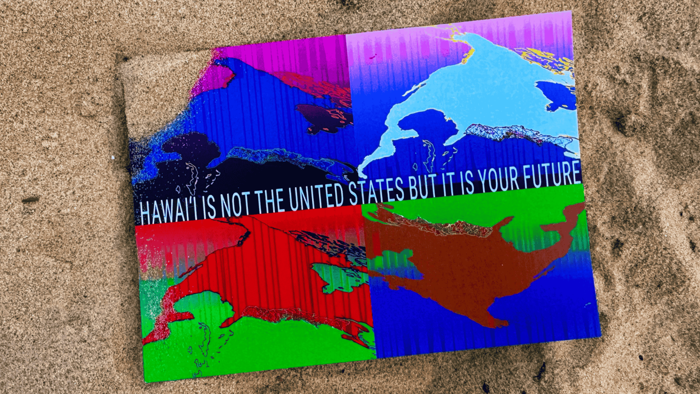 A photograph of a postcard laying in the sand. It reads "Hawai'i is not the United States, but it is your Future." It features multicolor maps of the east coast.