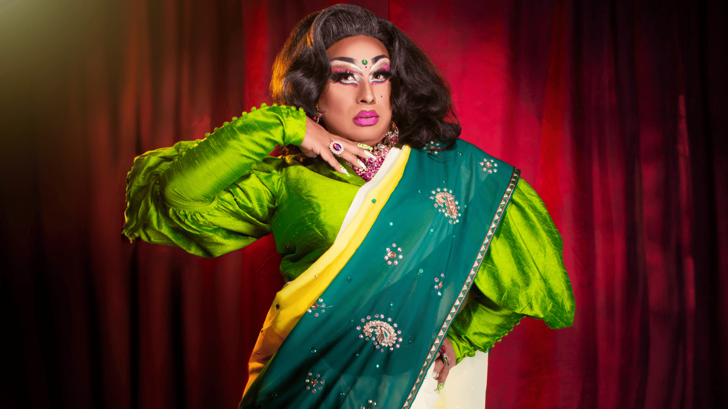 A photograph of Lawhore Vagistan. She wears a green and yellow sari and stands onstage in front of a red velvet curtain.