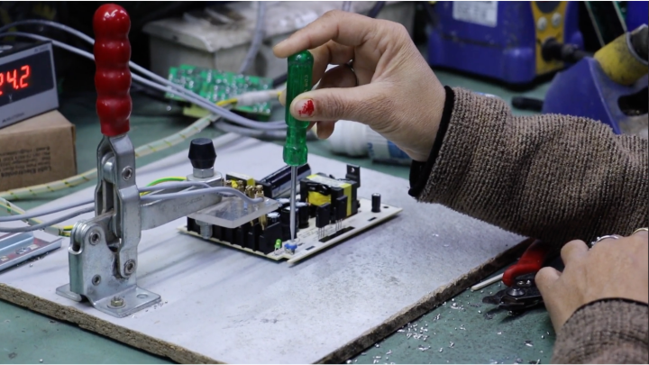 A photograph of a hand holding a screw driver above a circuit board in order to quality test it. The circuit board is placed on a factory table.