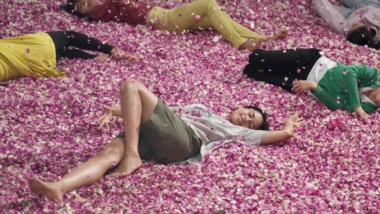 A still from the film Levitating. Characters from the film lie smiling in a bed of orchid petals, that also fall on them from above.