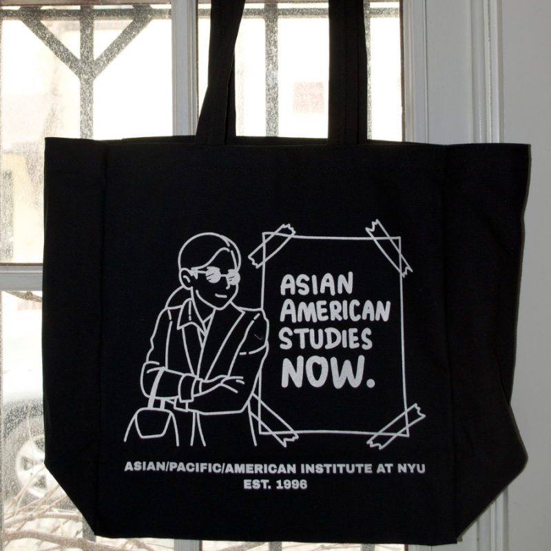 A photograph of a black tote bag with a white graphic image: a student stands near a sign that reads "Asian American Studies Now, Asian/Pacific/American Institute at NYU, EST. 1996"