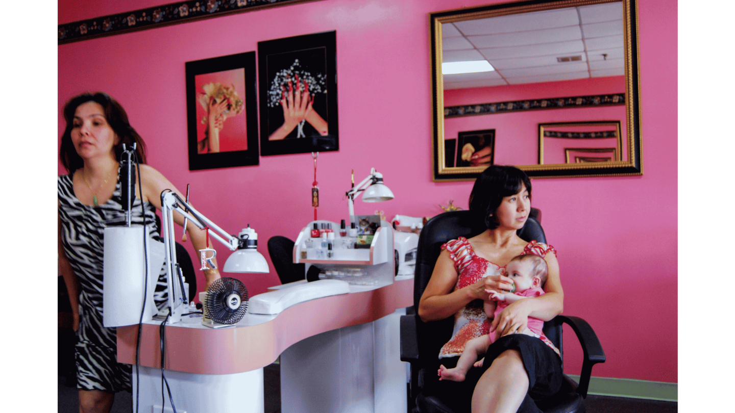 A photograph by Ocean Vuong features a nail salon worker feeding a baby as she sits in a salon chair.